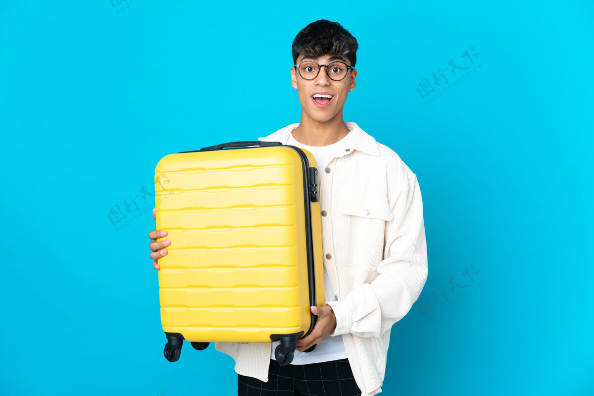 年轻年轻人在度假时带着旅行箱和蓝色的背景感到惊讶简短行李旅行