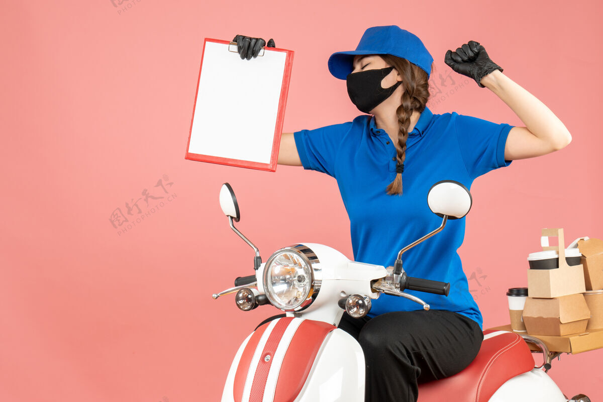 医疗前视图自豪的女快递员戴着医用口罩和手套坐在踏板车上 拿着空纸 在柔和的桃色背景下传递订单前面粉彩人