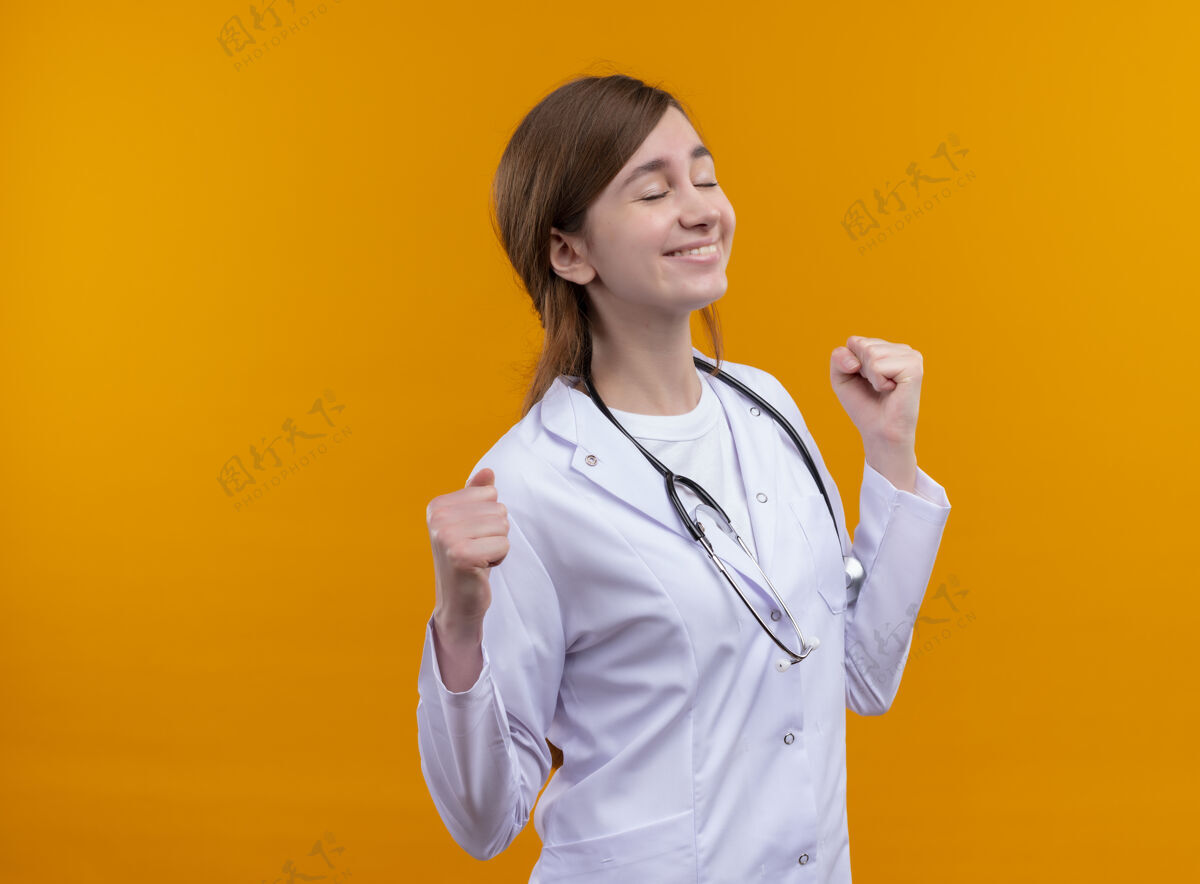 听诊器快乐的年轻女医生穿着医用长袍 手持听诊器 紧握拳头 闭上眼睛 在孤立的橙色空间与复印空间医生眼睛欢乐