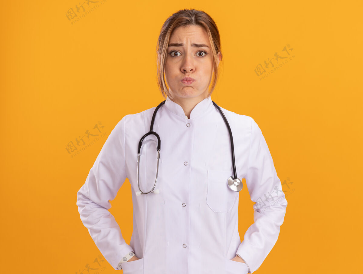 手年轻的女医生 脸颊浮肿 穿着医用长袍 手插在黄色墙上的口袋里长袍市民脸
