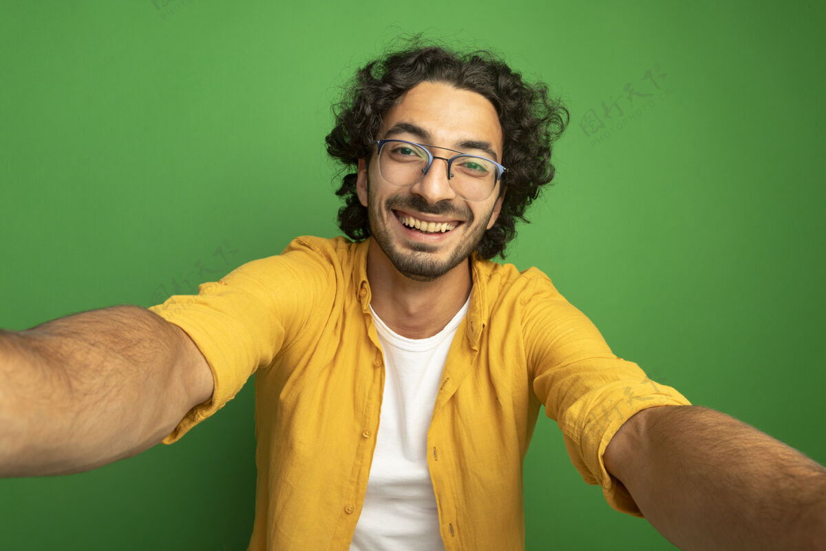眼镜快乐的年轻英俊的白种人戴着眼镜看着相机伸出双手对着隔离在绿色背景上的相机绿色走向快乐