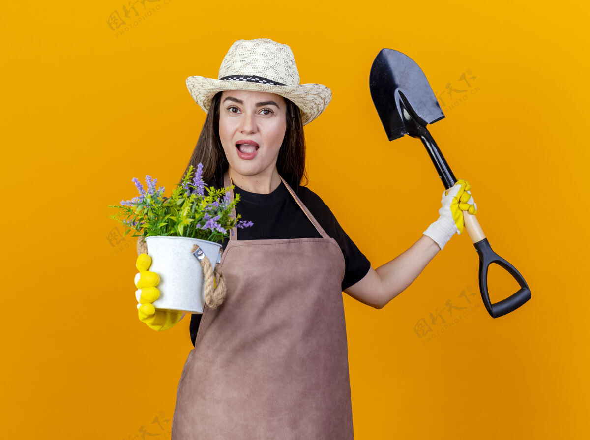 相机令人印象深刻的美丽的园丁女孩穿着制服 戴着园艺帽 戴着手套 手持铁锹 手持花盆里的花 在橙色背景上的镜头下隔离手握手套女孩
