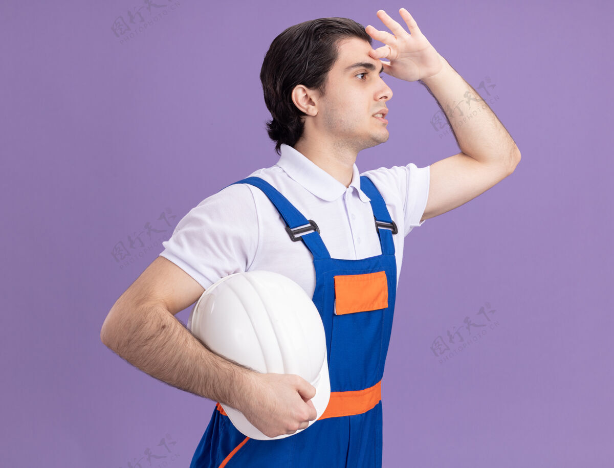 人身穿建筑制服的年轻建筑工人举着他的安全帽望着远处 手举着头站在紫色的墙上拿着制服头盔