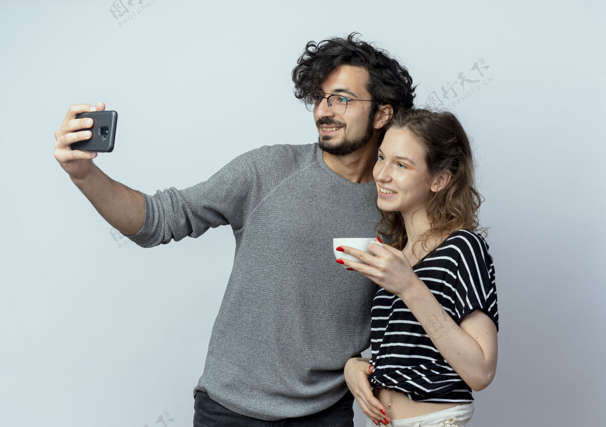 站立一对年轻的男女 快乐的男人用他的智能手机拍下了他们的照片 而他的女朋友站在他旁边喝着白色背景的咖啡使用情侣手机
