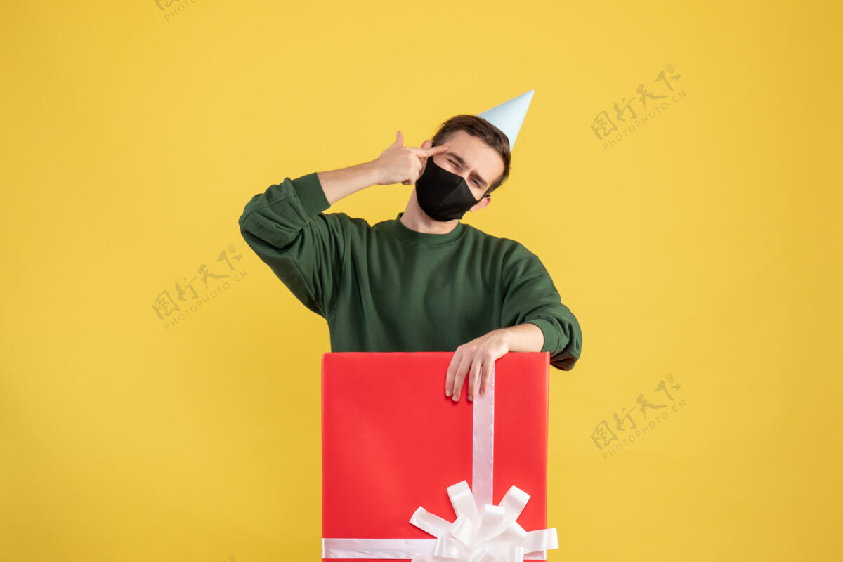 年轻人正面图：戴着派对帽和面具的年轻人站在黄色背景的大礼盒后面面具男性礼品盒