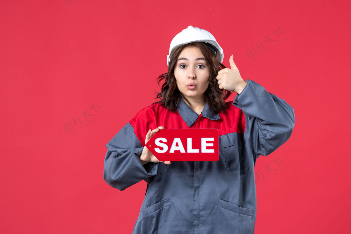 手势正面是穿着制服 戴着安全帽的好奇女工的近景 在隔离的红墙上展示销售图标并做出ok手势球衣吸引力衬衫