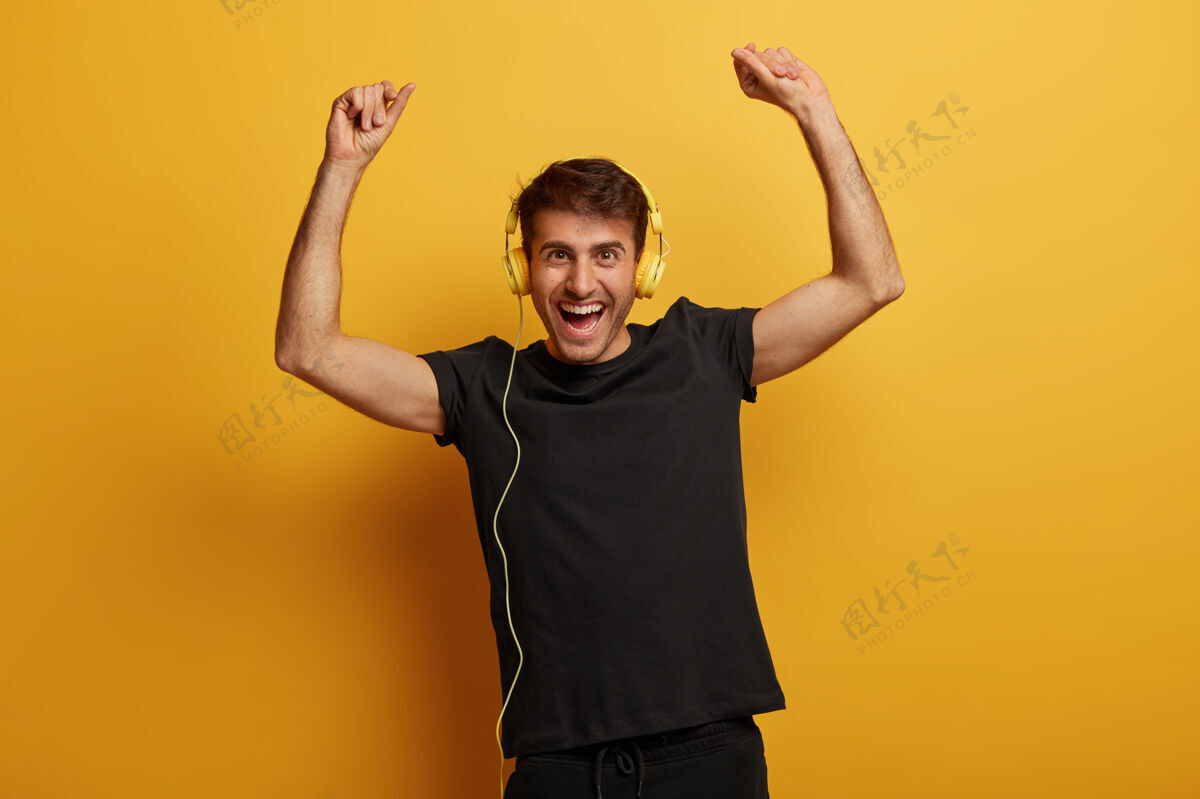 男性帅气精力充沛的男人幸福地举起双臂 戴着耳机 跟着最喜欢的歌唱歌 穿着黑色t恤 有着喜出望外的表情 孤立在黄色的背景之上快乐迪斯科声音