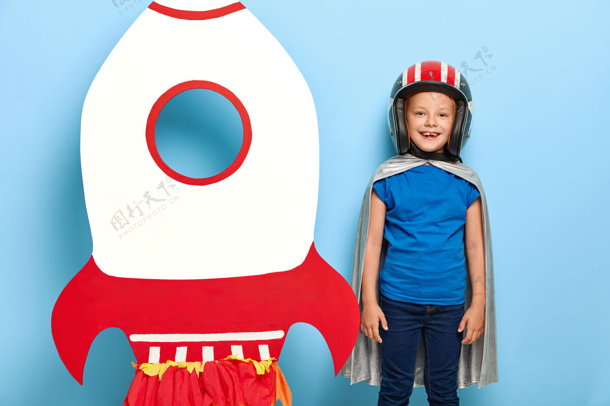 探索快乐的小孩戴着头盔和灰色斗篷 站在纸飞船火箭旁 想在太空中飞行服装头盔服装