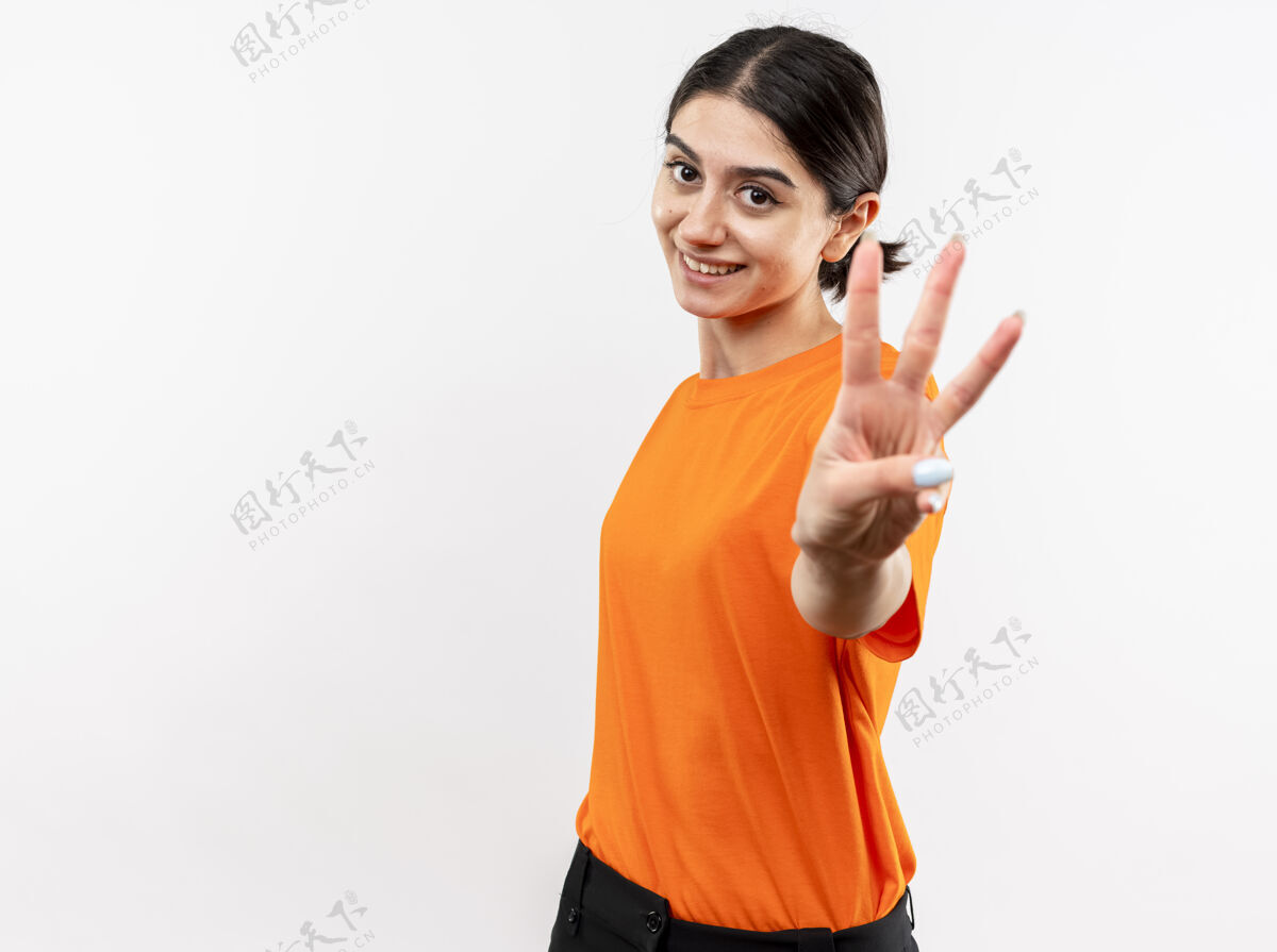 手指穿着橙色t恤的年轻女孩微笑着 手指着站在白墙上的三号手指年轻站数字