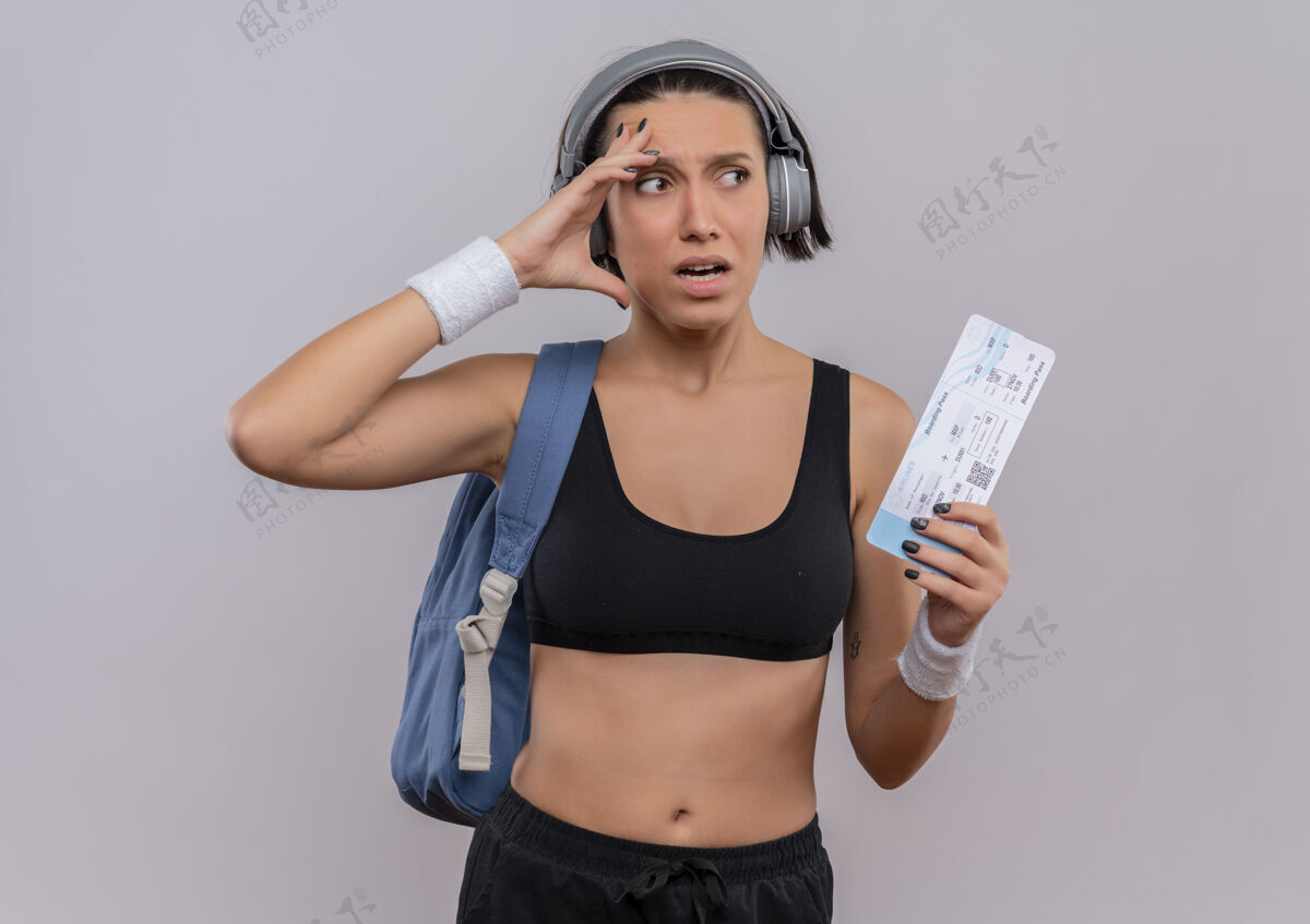 运动身穿运动服 头戴耳机 背着背包 拿着机票的年轻健身女士站在白色墙壁上困惑地看着一边 脸上露出恐惧的表情抱着头靠边站