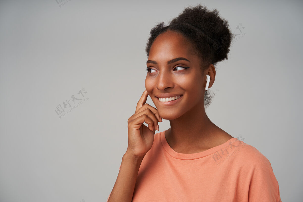 年轻年轻可爱的黑皮肤黑发女性的特写镜头 站在灰色的地面上 抬起手放在听筒上 开心地微笑着衣服耳机心情