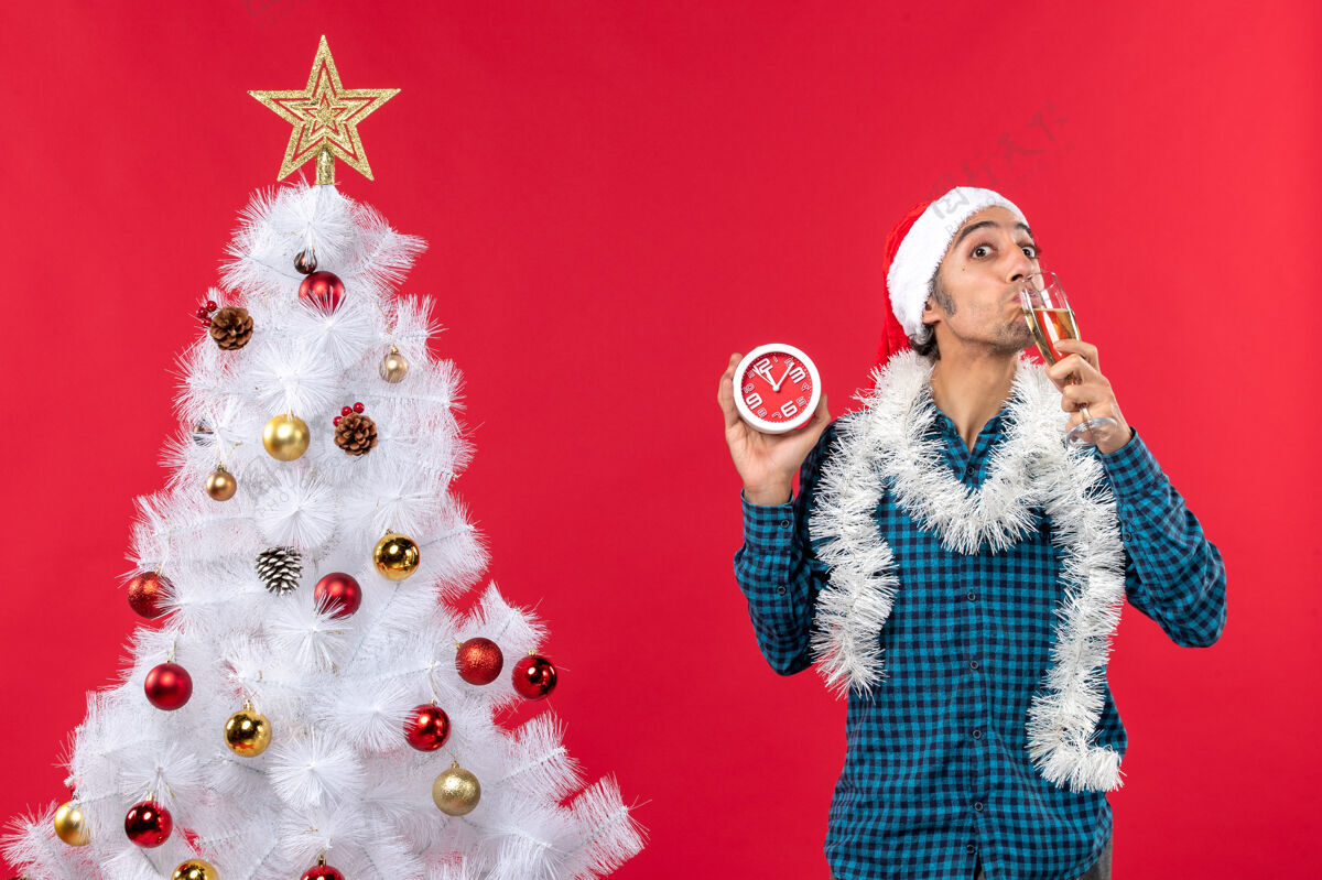 帽子带着圣诞老人帽子的微笑的年轻人 喝着一杯葡萄酒 拿着红色圣诞树旁的钟站着时钟庆典