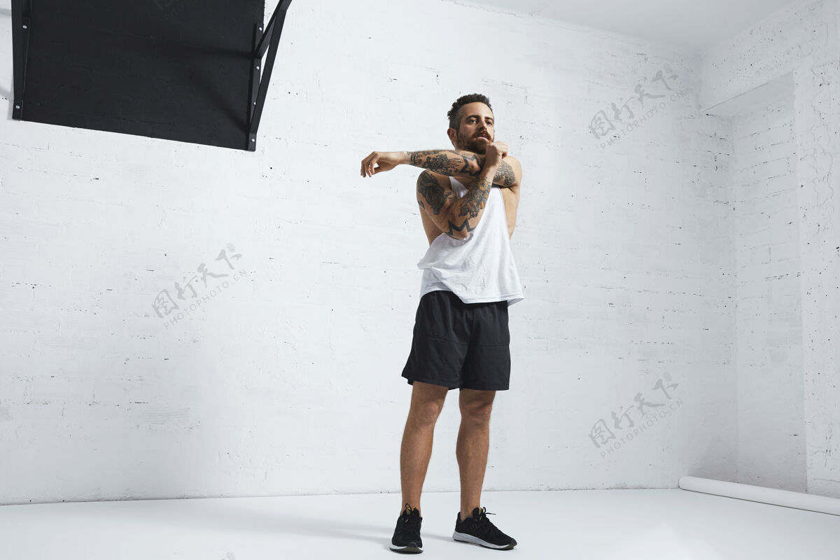 健康运动纹身男子在白色空白坦克t恤伸展他的肱二头肌在手臂上锻炼后 在砖墙旁边的黑色拉杆隔离健身房动作交叉