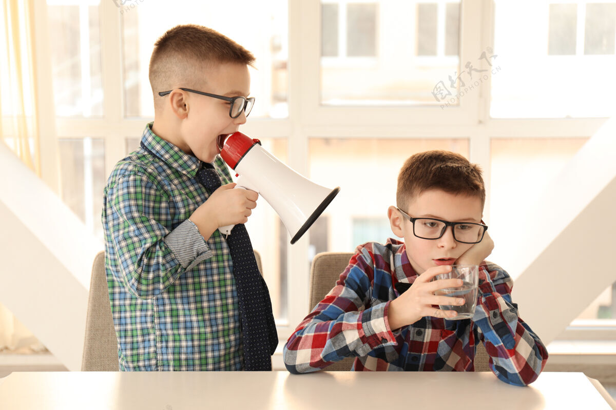 信息滑稽的小男孩拿着扩音器对着坐在桌边的无聊朋友大喊大叫信息情绪声音