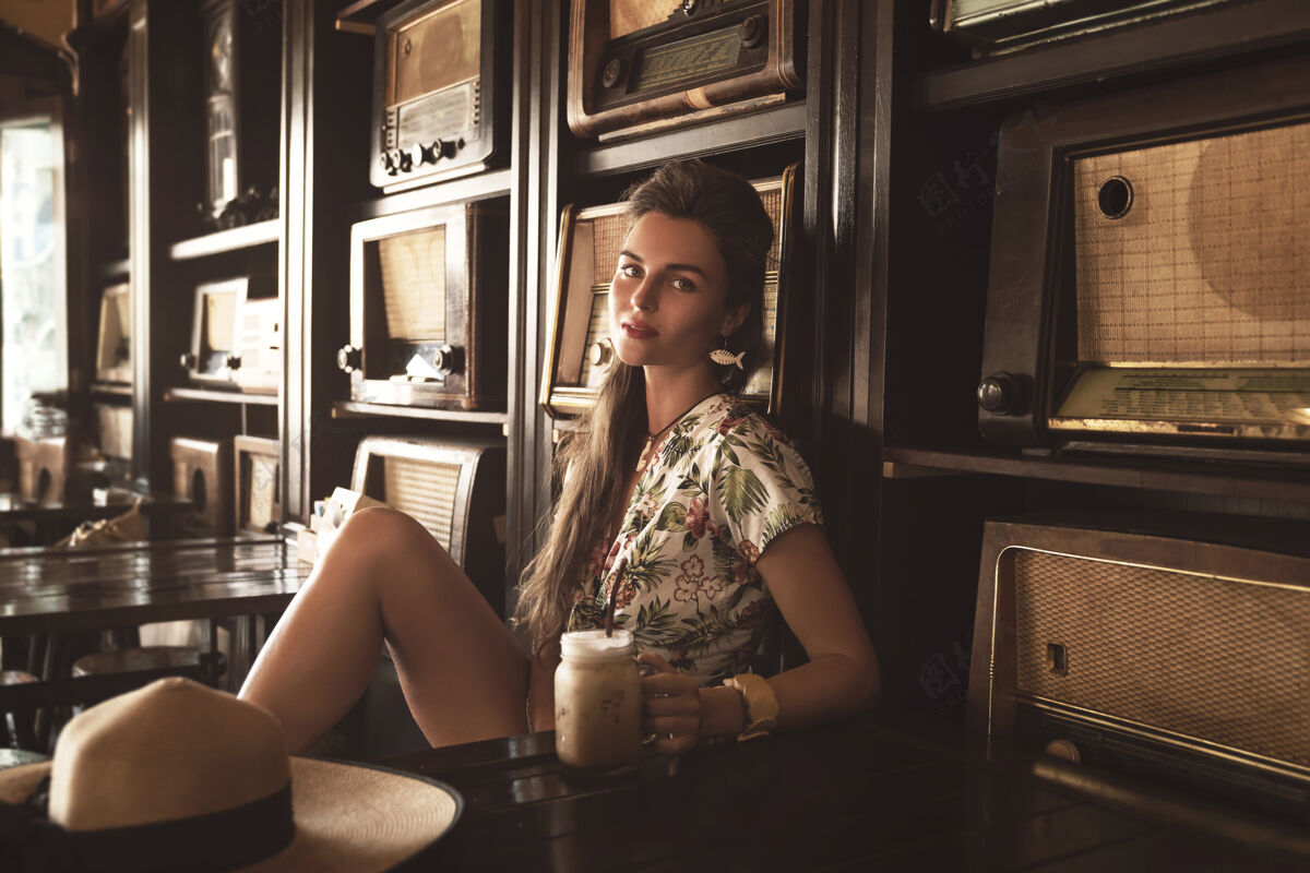 深褐色年轻快乐的女人在时尚复古的咖啡馆里喝着冰咖啡 里面有很多老式收音机模型收音机酒吧