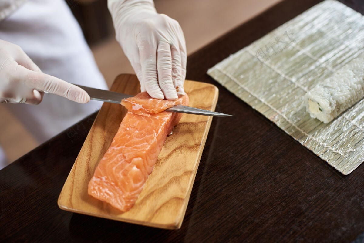 刀在餐厅准备美味的卷寿司过程的特写镜头女性手戴一次性手套在木板上切鲑鱼制作美味手