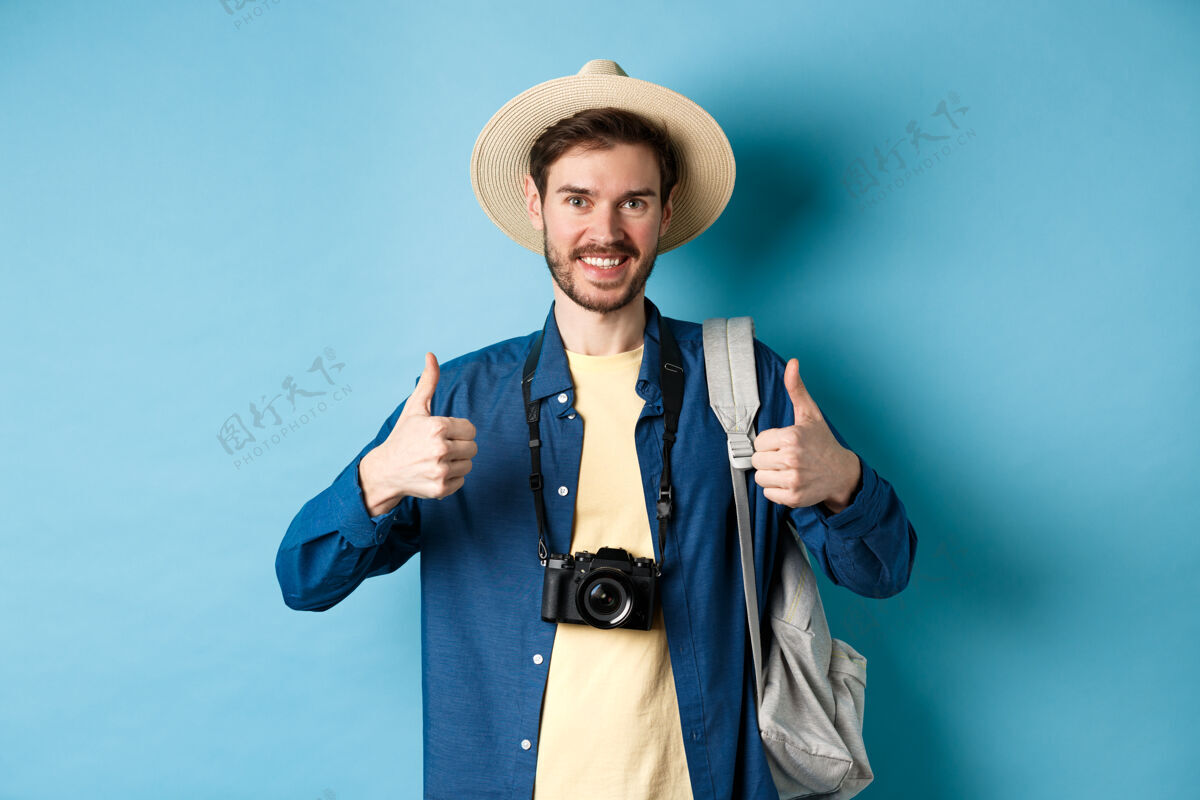 认可开朗帅哥推荐避暑胜地 竖起大拇指 微笑着表示赞许游客留下正面反馈 带着相机和背包站在蓝色背景上男自信推荐