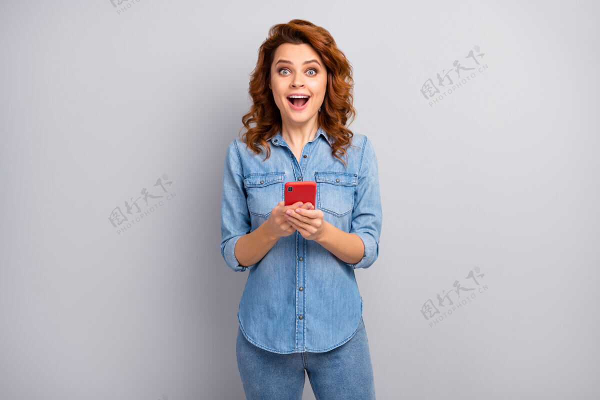 女孩惊讶的疯女人的肖像使用手机阅读社交媒体新奇印象深刻尖叫哇天哪穿好看的衣服隔离在灰色墙壁上新闻手机红发
