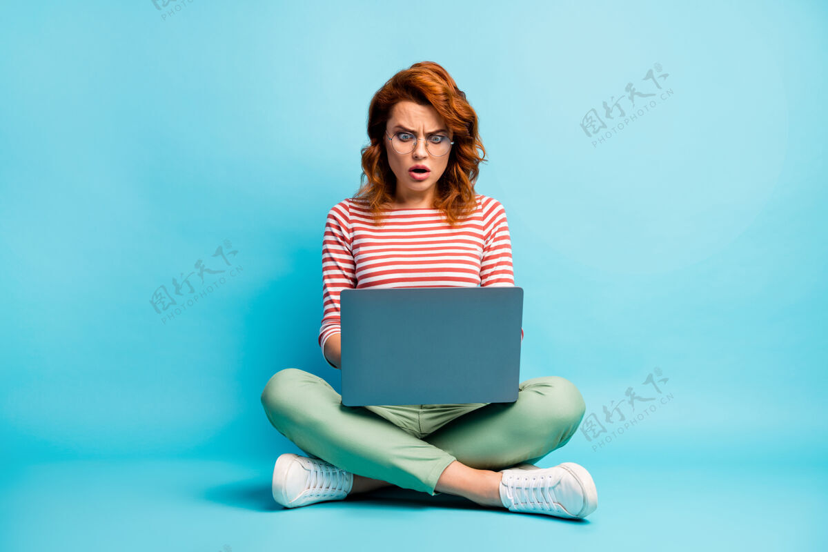 眼镜失意女孩的全身照片坐在交叉的工作电脑阅读社交媒体信息印象深刻尖叫难以置信意外穿时髦的套装鞋子孤立的蓝色同事红发休闲
