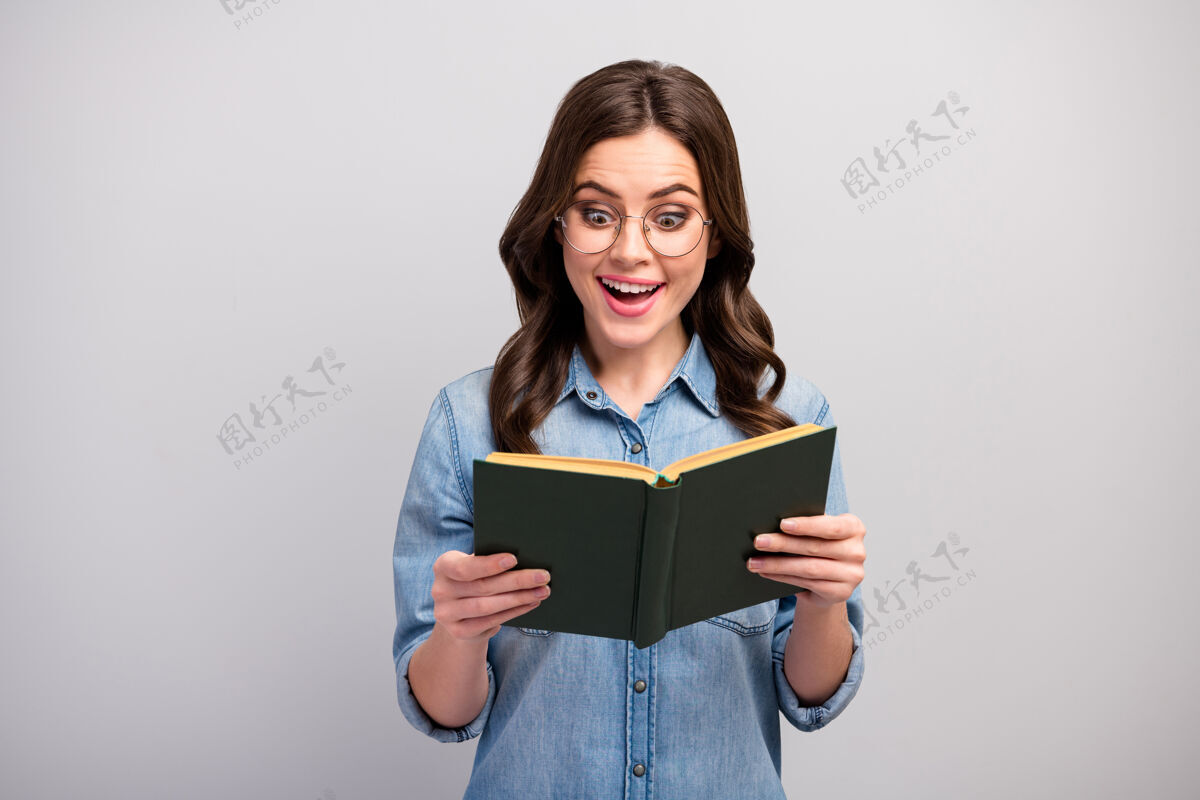 时尚照片中的俏皮搞笑的商务女士捧着书的手上瘾的读者书蠕虫惊人令人兴奋的快乐结局穿眼镜休闲牛仔裤牛仔衬衫孤立的灰色Omg年轻眼镜
