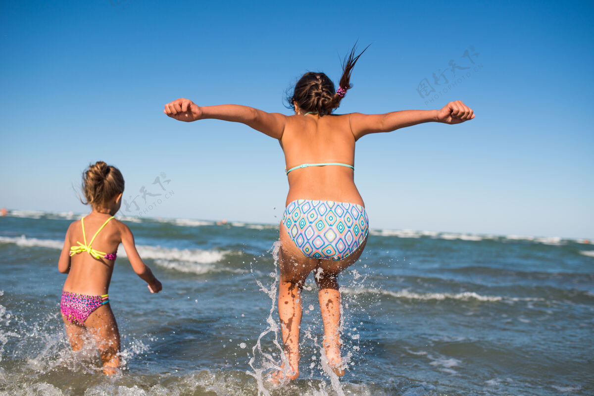海滩两个有趣的小女孩在喧闹的海浪中跳跃 在一个阳光明媚的夏日享受期待已久的假期跳跃万里无云风