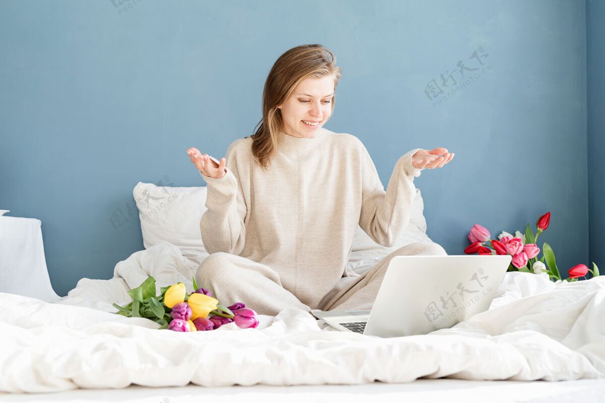 漂亮快乐微笑的女人穿着睡衣坐在床上 愉快地赏花 用笔记本电脑聊天年轻庆祝微笑