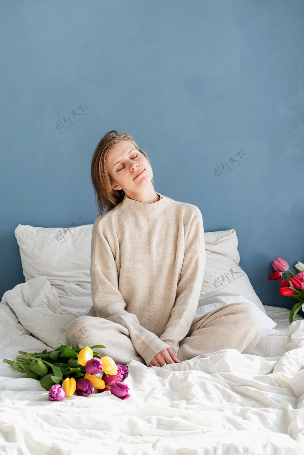 浪漫快乐的女人穿着睡衣坐在床上捧着郁金香花束 背景是蓝色的墙壁生活方式美丽节日