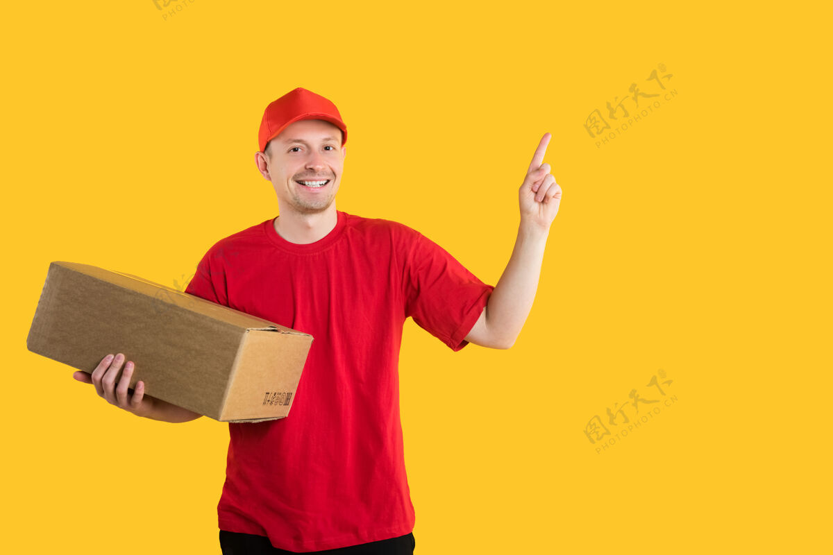 送货穿着红色制服的快乐信使拿着一个科尔顿盒子 用手指着黄色上面的自由空间纸板快递帽子