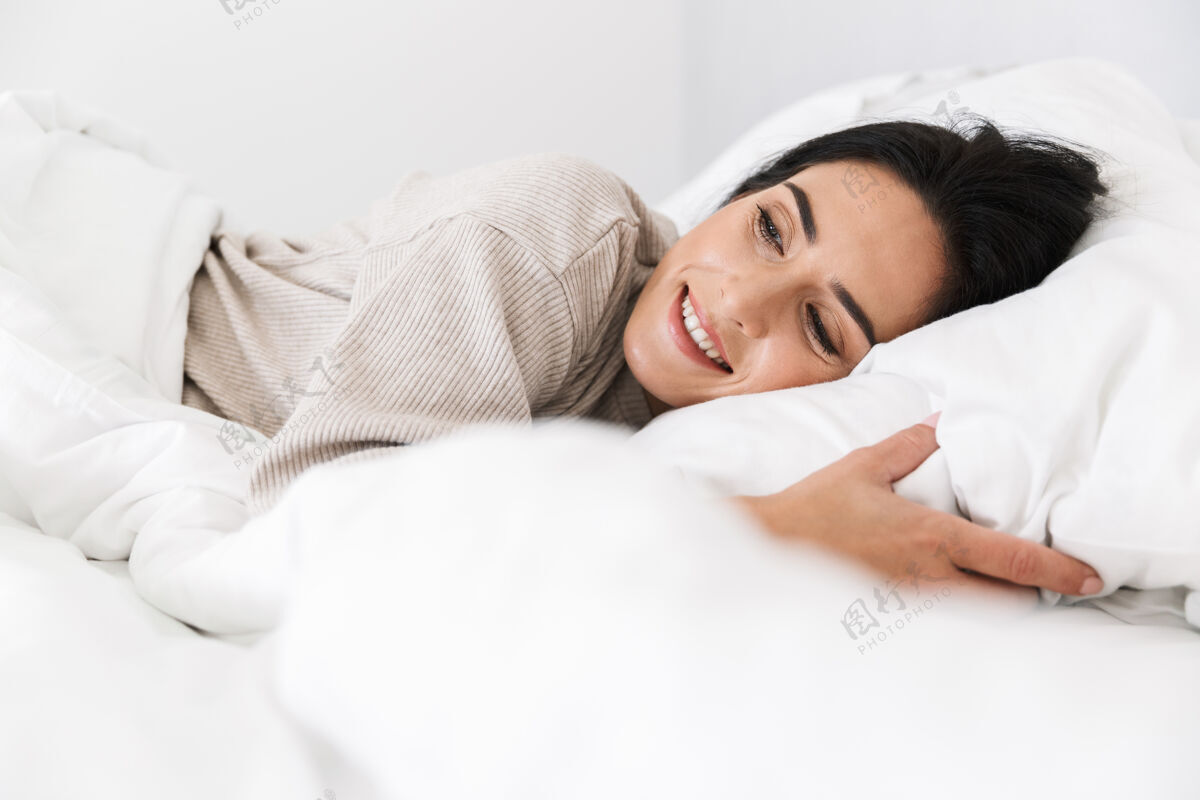 深色头发可爱的女人30多岁的照片微笑 而躺在床上与白色亚麻布在家里放松愉快亚麻布
