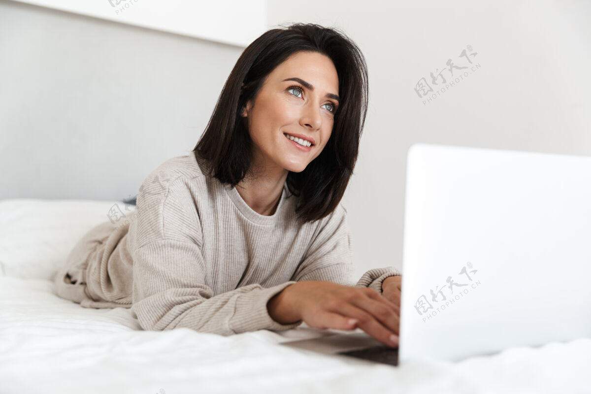 舒适可爱的女人30多岁的照片 使用笔记本电脑 而躺在床上与白色亚麻布在明亮的房间中年吸引力室内
