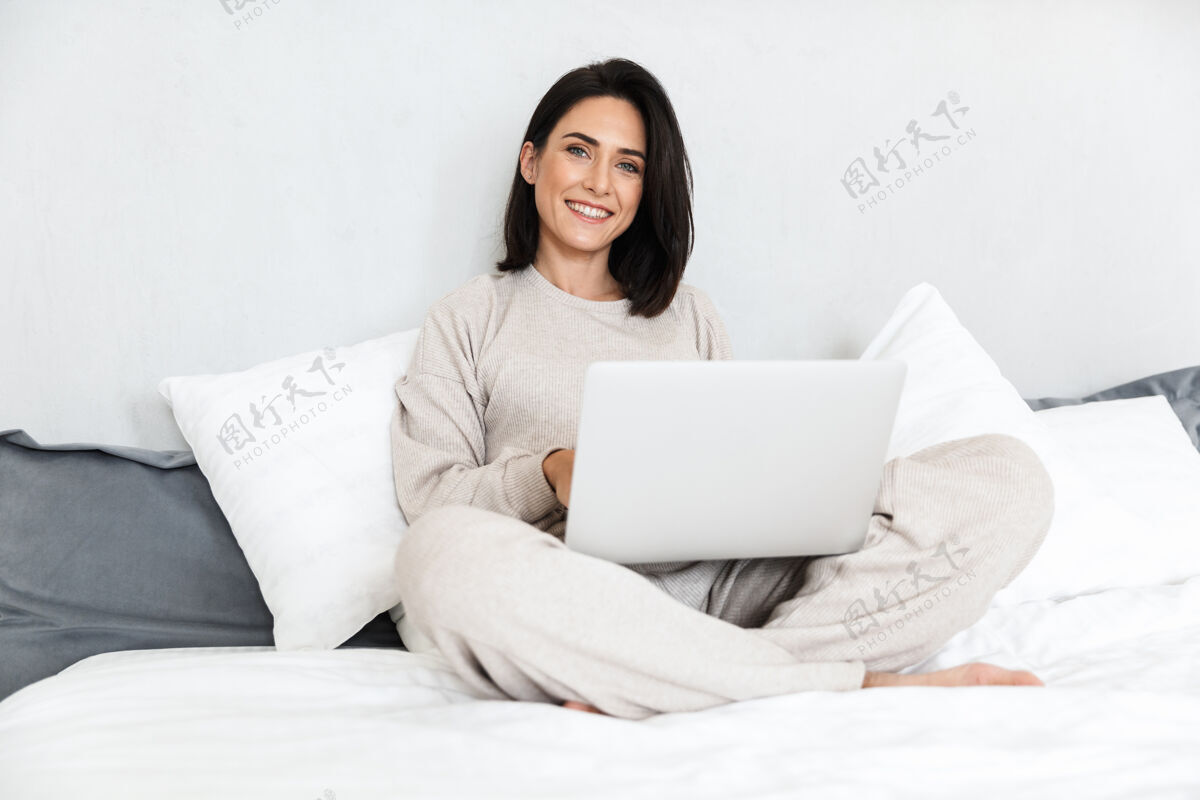 公寓一张30多岁的快乐女人在舒适的公寓里用笔记本电脑 穿着白色亚麻布坐在床上的照片舒适女士中年