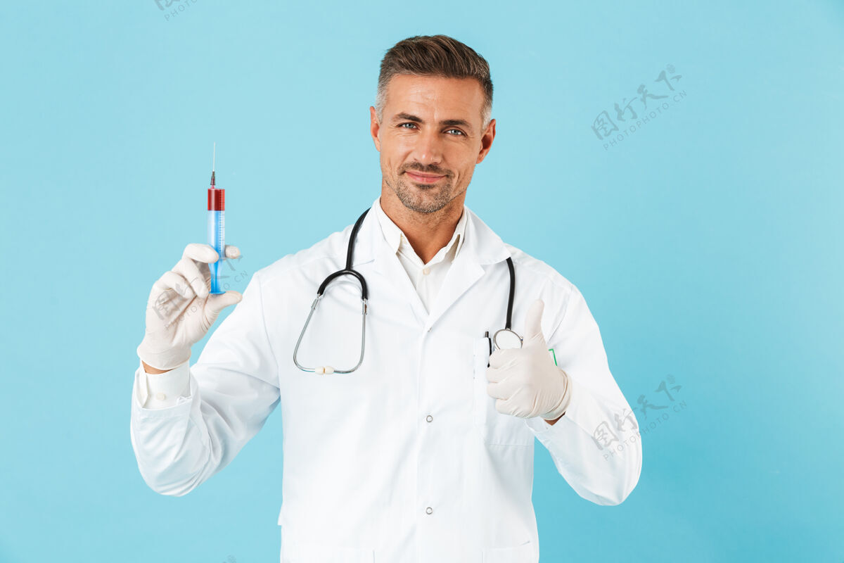 医疗戴着听诊器戴着手套拿着注射器的白人医生的画像 孤立地站在蓝色的墙上专家成人听诊器