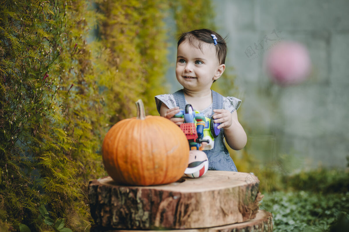 树可爱有趣的女婴站在自家院子里装饰性的脚踩和南瓜旁边 面带微笑生活方式公园高加索