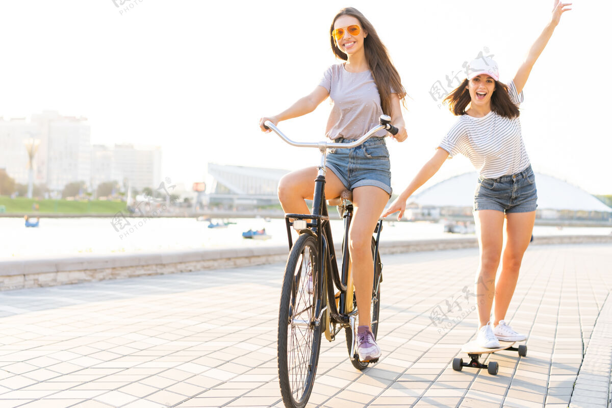 火炬两个年轻的朋友在一个炎热的夏日放松 一个年轻的女人骑着自行车拖着她的朋友在一条城市街道上滑滑板自行车玩城市