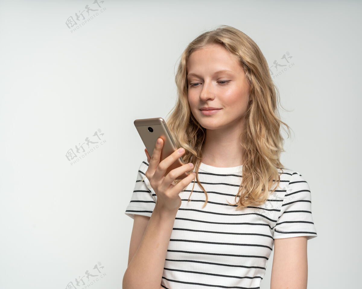 打字站在白墙附近 灰色背景下 漂亮的女性在手机上查看社交媒体长发金发聊天