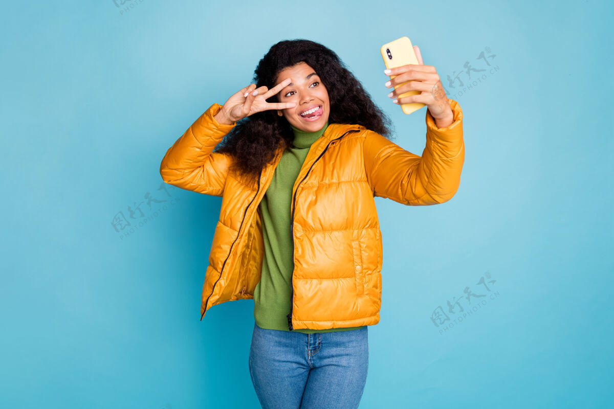 多民族时髦的深色皮肤女士拿着电话自拍秀v字贴舌头露嘴穿时髦的黄色秋季外套牛仔裤绿色毛衣蓝色隔离墙多国风衣混合
