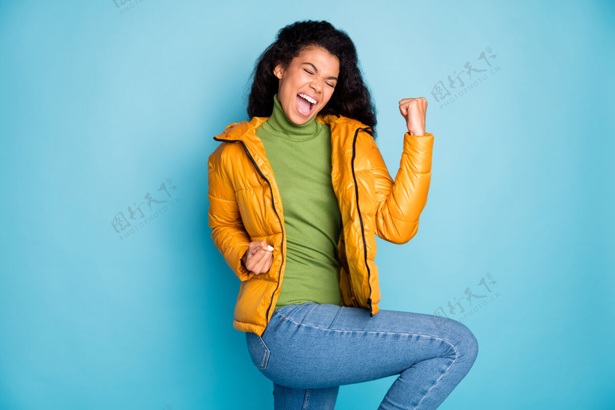 牛仔裤惊人的深色皮肤卷曲女士庆祝足球比赛的目标尖叫穿时髦的黄色春季大衣牛仔裤绿色套头衫孤立的蓝色墙壁毛衣欢乐年轻