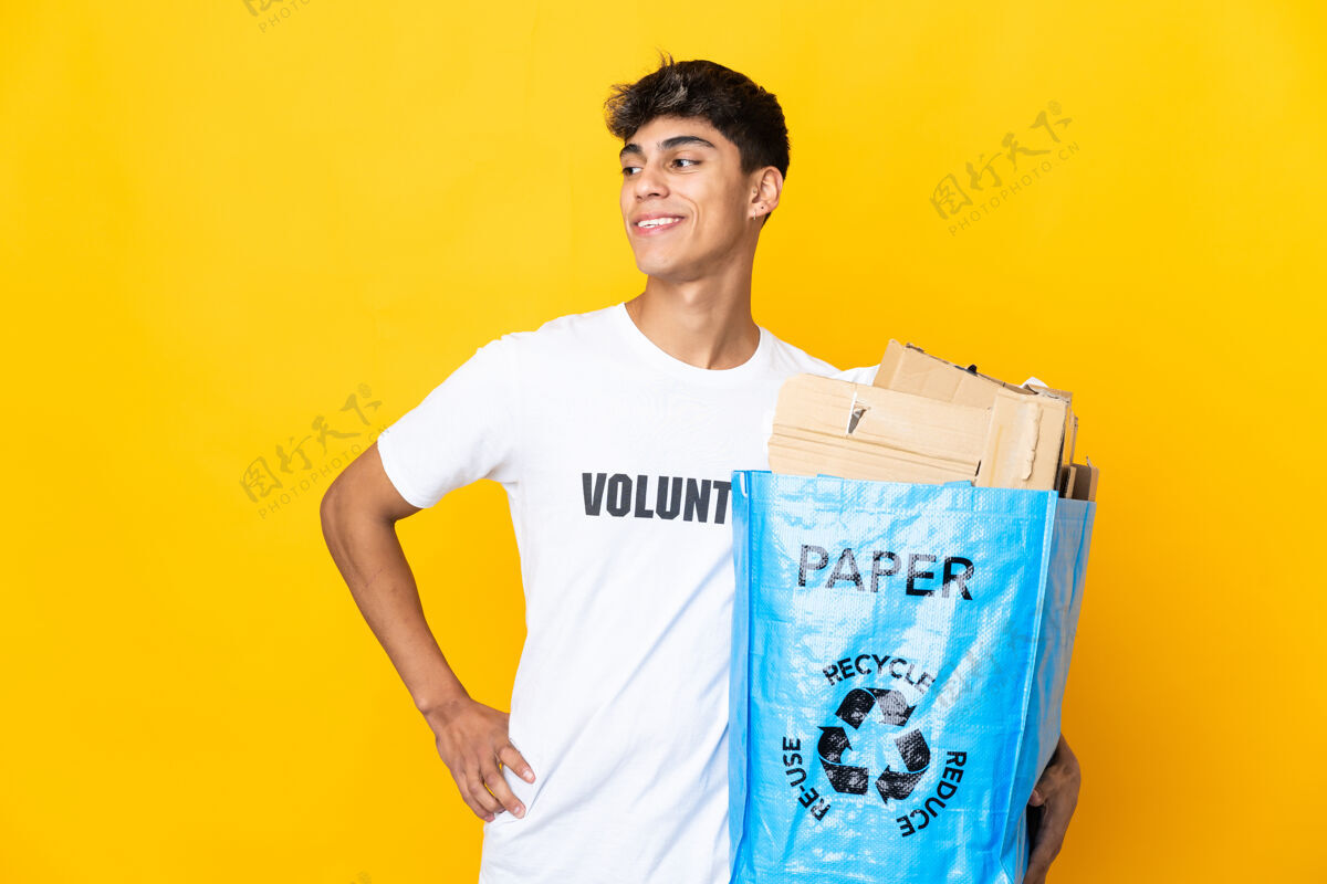 人一名男子拿着一个装满废纸的回收袋在隔离的黄色墙壁上进行回收 双臂放在臀部 面带微笑手臂责任垃圾