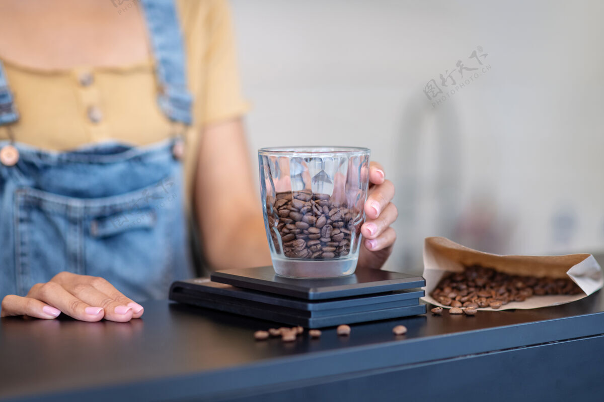 咖啡豆全豆咖啡女士手把一个装有咖啡豆的玻璃杯放在柜台的秤上 看不到一张脸准确香气职业