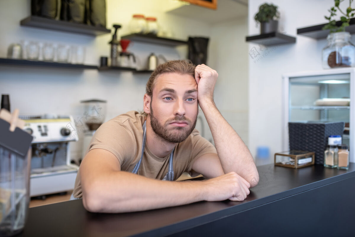 沮丧失望沮丧一个穿着条纹围裙和T恤的年轻人坐在咖啡馆的吧台后面 无聊极了围裙启动自营职业