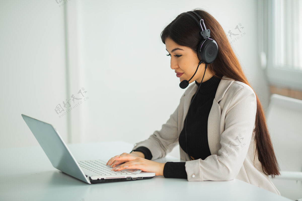 耳机一个穿着西装的漂亮年轻女子坐在办公室的桌子旁 手提电脑插着耳机互联网中心工作