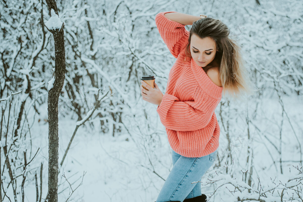 享受一个穿着粉色宽松毛衣和牛仔裤 手里拿着咖啡的漂亮女孩 在寒冷的雪域森林里寒冷靴子公园