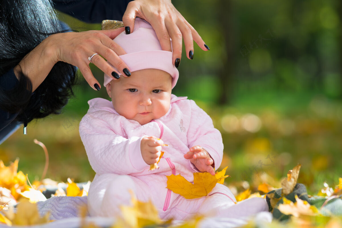 枫树妈妈把一顶粉红色的帽子戴在她的小女儿的头上 她坐在公园的地毯上玩黄色的秋叶婴儿秋天婴儿