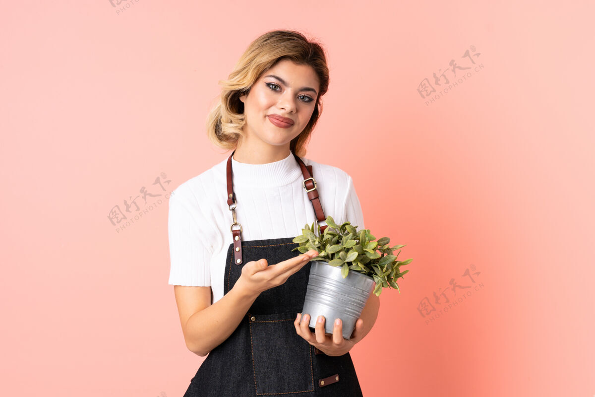 花盆俄罗斯园丁女孩手捧一株孤立的粉红色植物向一旁伸出双手 邀请前来金发呼叫朋友