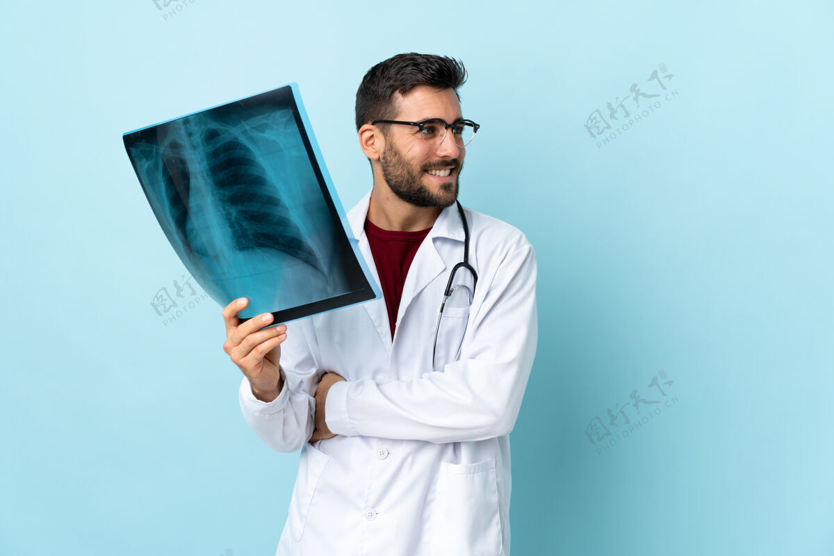 胡须专业的创伤科医生拿着隔离在蓝色背景上的X光片往侧面看男人快乐年轻