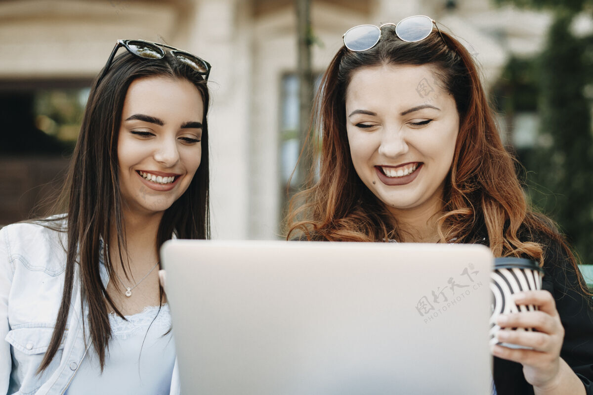 城市两个可爱的年轻女朋友一边看着笔记本电脑屏幕一边喝着咖啡 一边坐在靠着一栋楼的长凳上笑着放松约会美丽