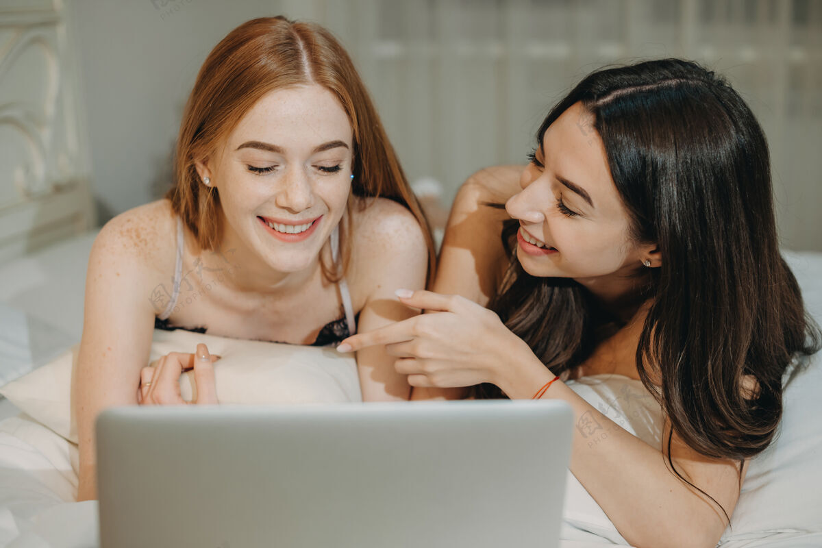 空闲时间两个可爱的女朋友坐在床上笑着看着笔记本电脑屏幕的特写镜头在线床肖像