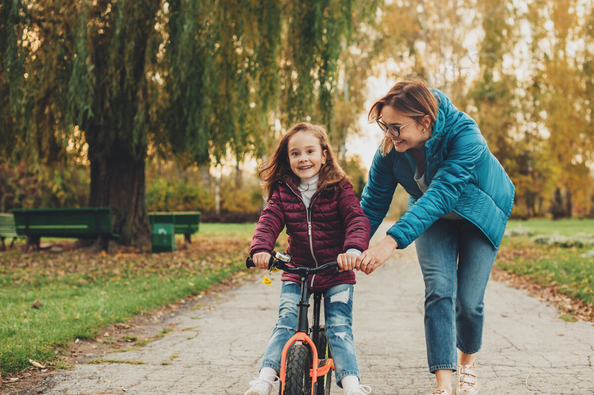 情感美丽的小女孩在公园里骑自行车 而她的妈妈在户外帮忙关系学习女人