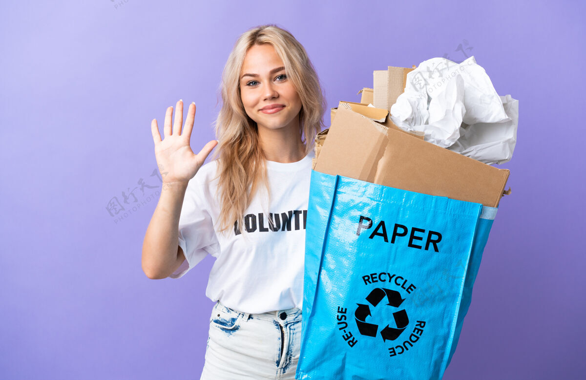 金发年轻的俄罗斯女子拿着一个装满废纸的回收袋 在紫色的背景上用手指数着5个回收手想法