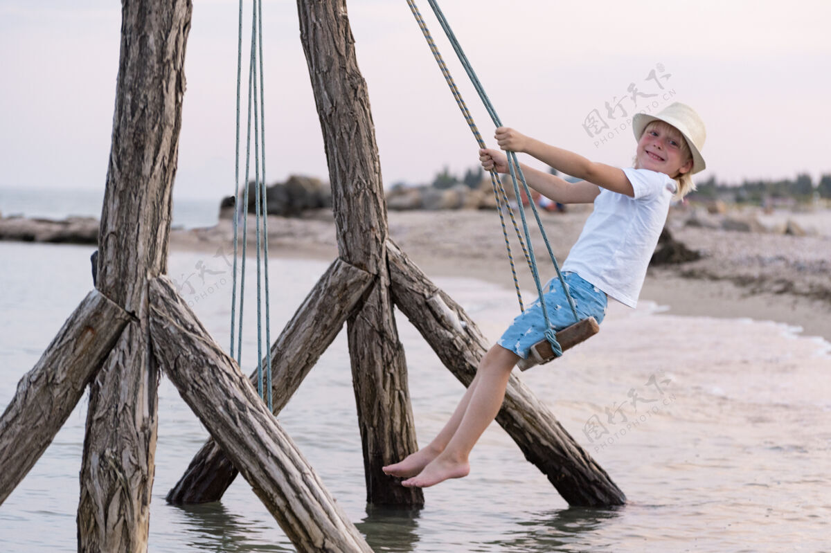 男孩快乐的孩子骑在绳子上荡秋千水秋千由海上假日孩子们在海上海岸度假休息